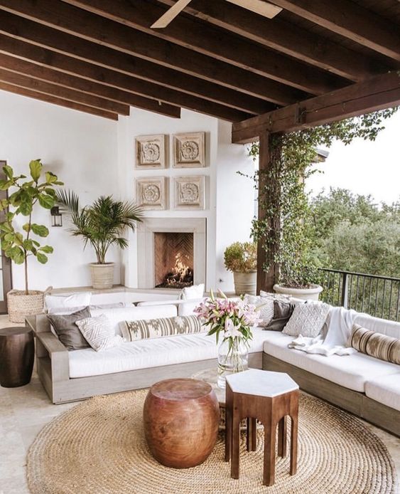 a neutral boho living room with a fireplace, a corner sofa with printed pillows, wood and metal tables and potted greenery
