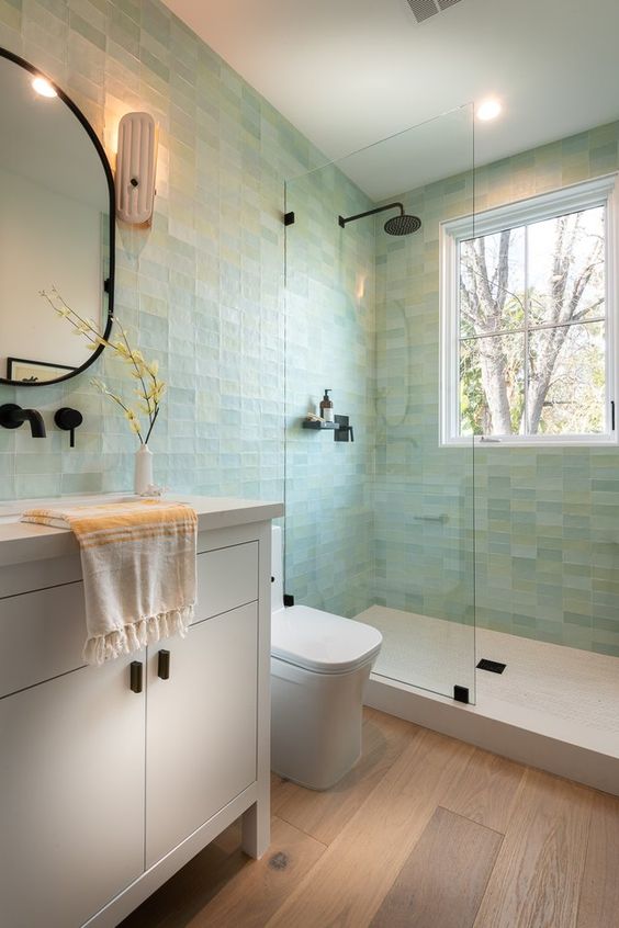 a beautiful modern bathroom clad with pastel green tiles, a white shower, vanity and appliances, black fixtures
