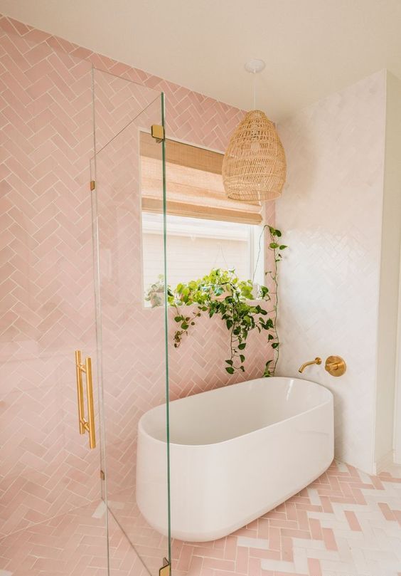 a beautiful pink and white bathroom with herringbone tiles, an oval tub, gold fixtures and a woven pendant lamp