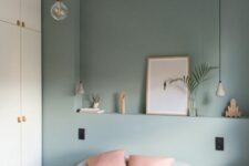 a bedroom with a pastel green accent wlal, a bed with grey and pink bedding, a white wardrobe and pendant bulbs