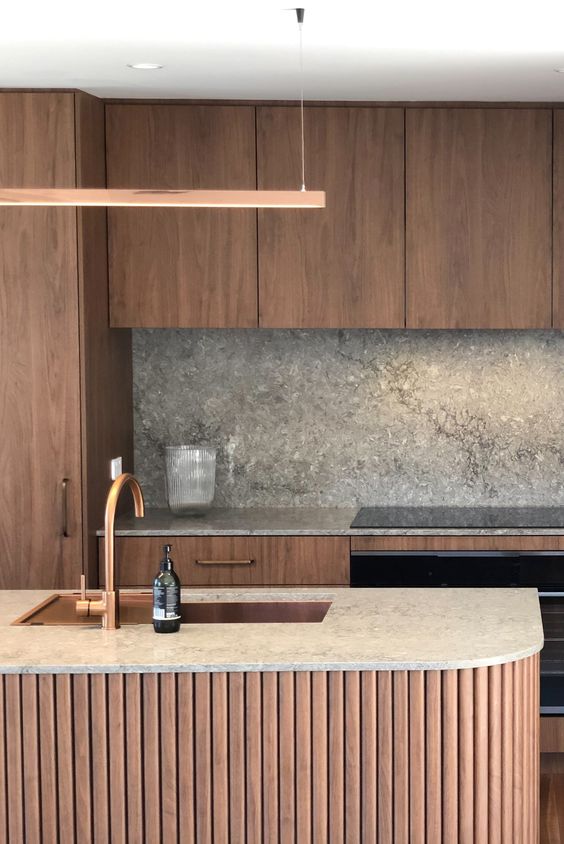 a chic and cozy kitchen with stained cabinets, a fluted curved kitchen island and grey stone countertops and a backsplash