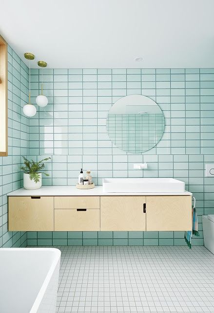 a chic bathroom with light blue and white tiles, a floating vanity, a tub, a round mirror and potted plants
