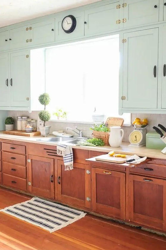 a chic vintage farmhouse kitchen with stained and light green cabinets, neutral countertops, potted greenery and printed textiles