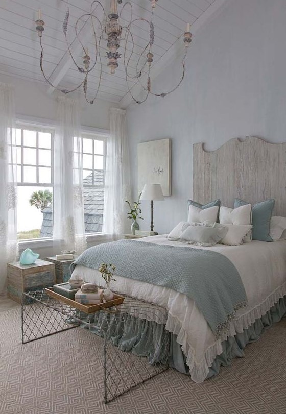 a coastal shabby chic bedroom with a wooden bed with a statement headboard, blue and white bedding, wooden furniture, a crystal chandelier and a wire bench