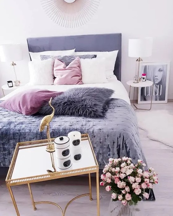 a cool feminine bedroom with a purple bed, a gold nightstand with candles, pink blooms, pink, white and purple pillows