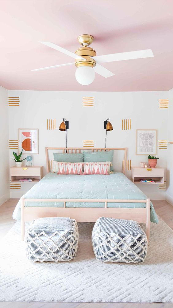 a cool pastel bedroom with a pink ceiling, a light stained bed and nightstands, pastel bedding, blue and white poufs