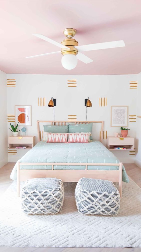 a cool pastel bedroom with a pink ceiling, a light-stained bed and nightstands, pastel bedding, blue and white poufs