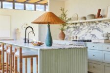 a cozy and welcoming mint green kitchen with shaker style cabinets, an open shelf instead of upper ones, a white marble backsplash, a fluted kitchen island