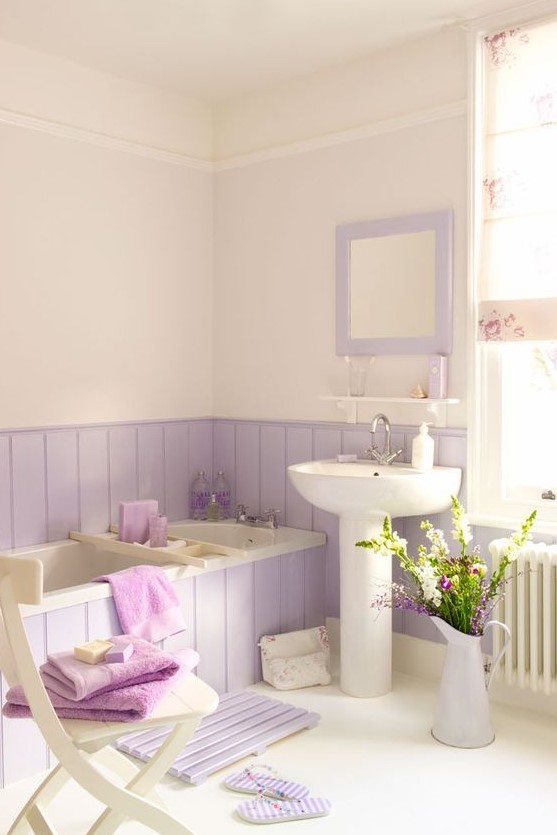 a cute and lovely bathroom with lilac paneling around the tub and a mirror and a mat of matchine shades, with blooms and a chair