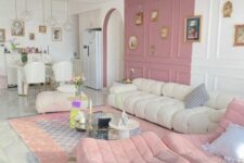 a delicate living room with a color block pink and white wall, a creamy low sofa, pink ottomans, neutral tables