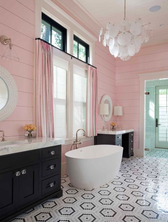 a delightful bathroom with pink slatted walls, black vanities, an oval tub, a geo tiled floor and a scale chandelier