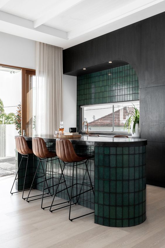 a dramatic kitchen with black cabinets and a curved kitchen island clad with green skinny tiles plus a matching wall