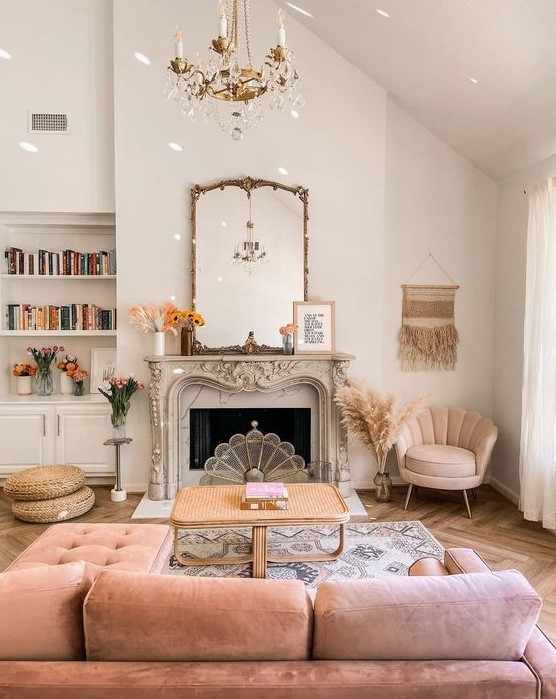 a dreamy living room with a vintage fireplace, a pink sectional and a pink chair, a cane coffee table, a gold chandelier plus books