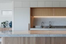 a large contemporary kitchen with lit up upper cabinets, a tile backsplash, a ridged kitchen island with a grey stone countertop