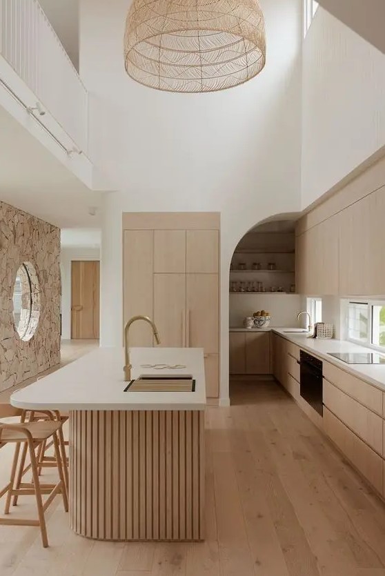 a light stained minimalist kitchen with sleek cabinets, a window backsplash, a curved fluted kitchen island with a curved part and a woven pendant lamp