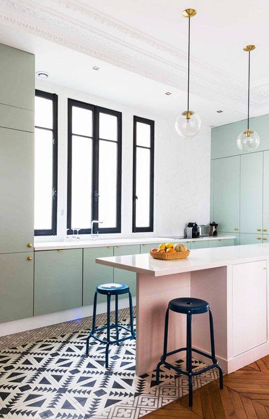 a lovely pastel kitchen with pastel green cabinets and a pink kitchen island, black and white tiles and black stools