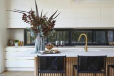 a minimalist white kitchen with a window backsplash, a large fluted kitchen island with black stools and gold touches here and there