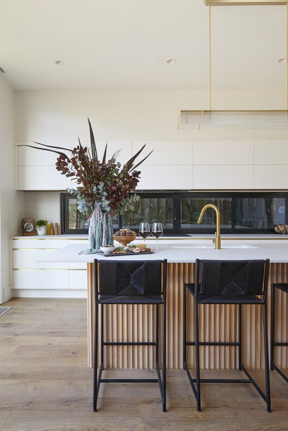 a minimalist white kitchen with a window backsplash, a large fluted kitchen island with black stools and gold touches here and there
