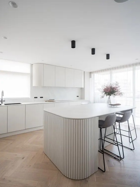a minimalist white kitchen with curved cabinets, a white marble backsplash, a curved ribbed kitchen island and tall grey stools