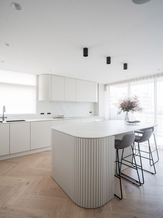 a minimalist white kitchen with curved cabinets, a white marble backsplash, a curved ribbed kitchen island and tall grey stools