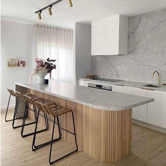 a minimalist white kitchen with fluted cabinets, a white marble backsplash, a curved kitchen island with a stone countertop