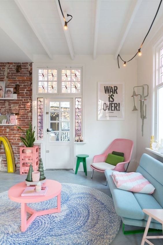 a modern and fresh pastel living room with a brick wall, a pink chair, stand and a round table, a blue sofa, a blue rug and some pillows