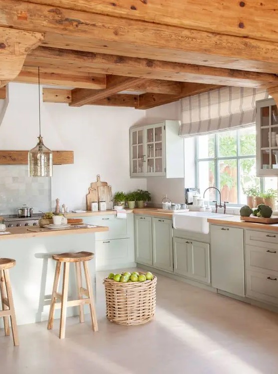 a modern cottage kitchen with wooden beams on the ceiling, light green shaker style cabinets, butcherblock countertops, a basket for storage and a pendant lamp