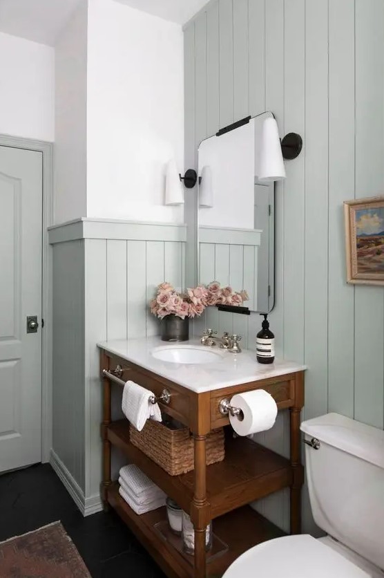 a modern country bathroom with pale aqua paneling, a stained vanity, white appliances, vintage lamps and some blooms