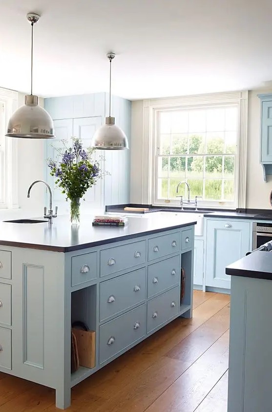 a modern country light blue  kitchen with dark countertops, pendant lamps and large windows plus a lovely hardwood floor is chic