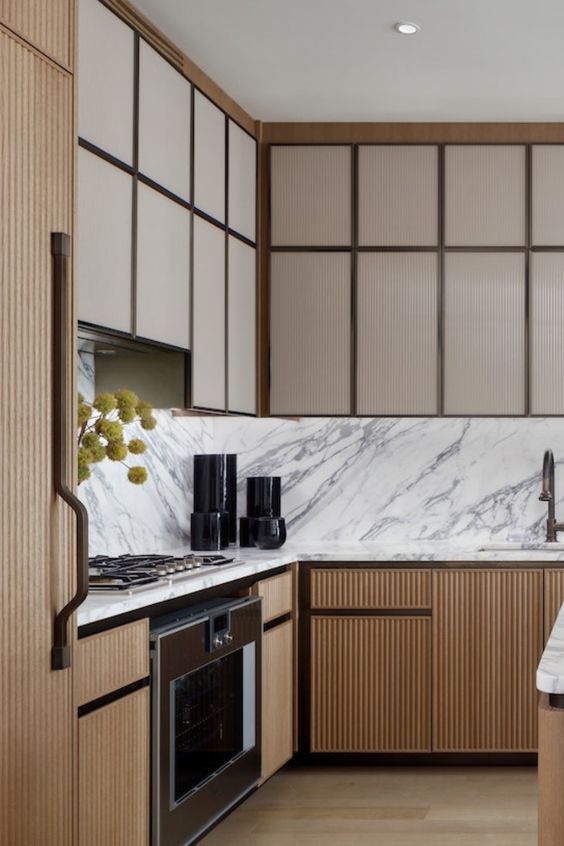 a modern kitchen with beige and stained fluted cabinets, a white marble backsplash and countertops plus black appliances