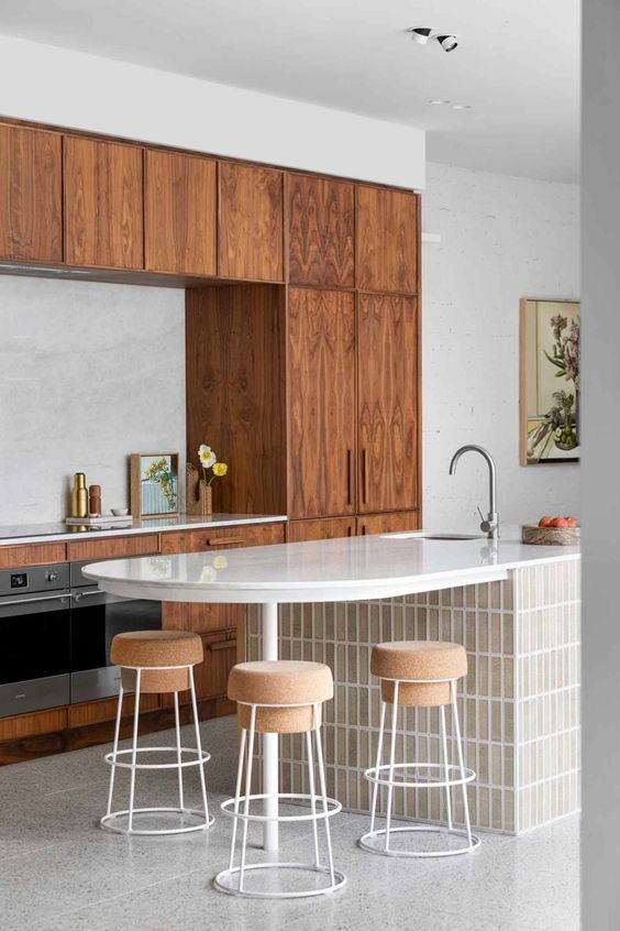 a modern kitchen with stained cabinets, a curved kitchen island clad with skinny tiles, cork stools and a white stone backsplash