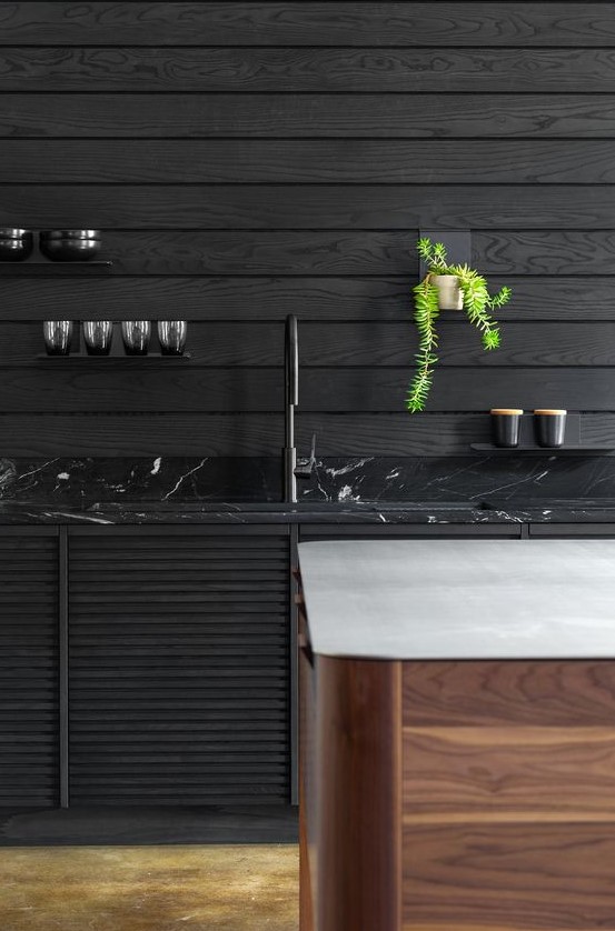 a moody kitchen with black plank walls, a black marble countertop and a wooden kitchen island with a white stone countertop