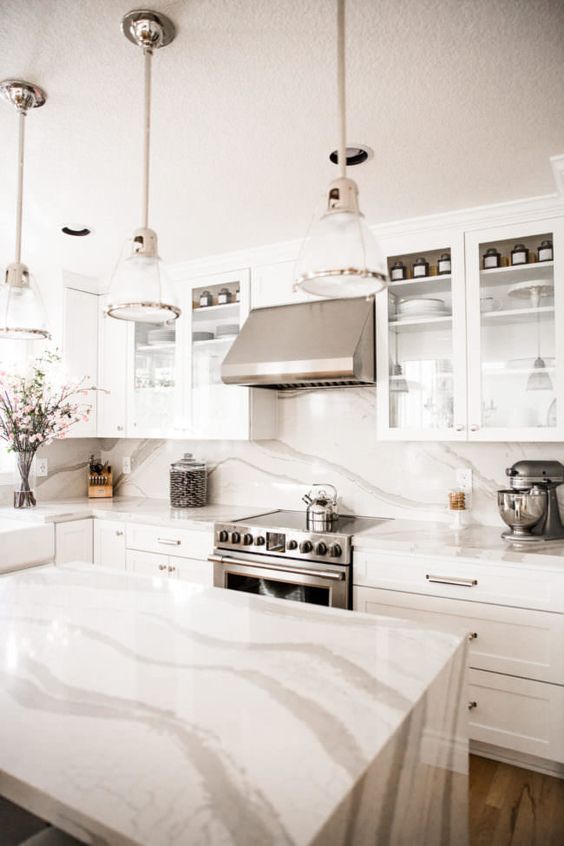 a neutral kitchen with shaker cabinets and glass ones, a white quartz backsplash and matching countertops, elegant lamps