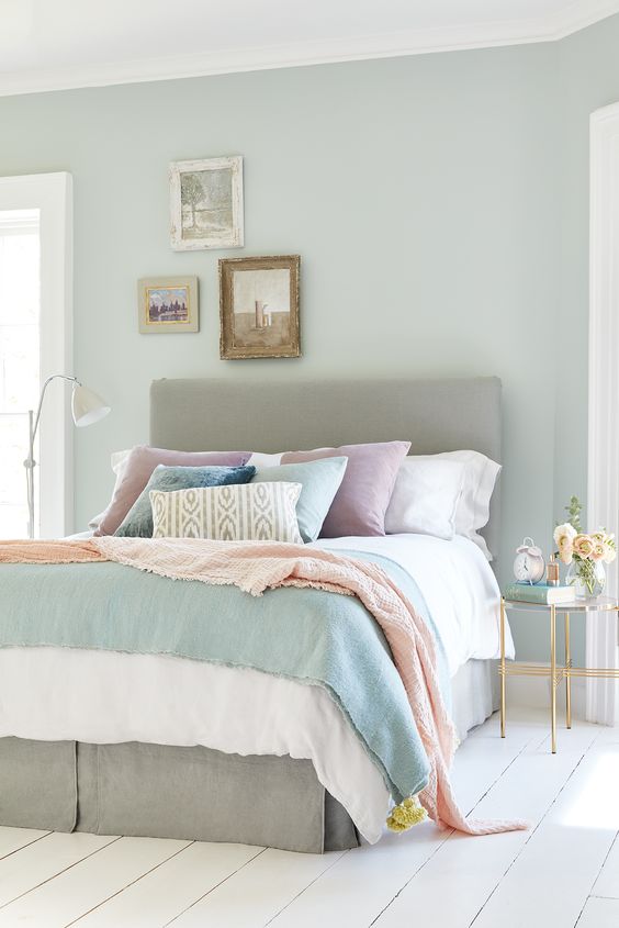 a pastel bedroom with a grey upholstered bed with pastel bedding, a mini gallery wall, a refined nightstand with blooms