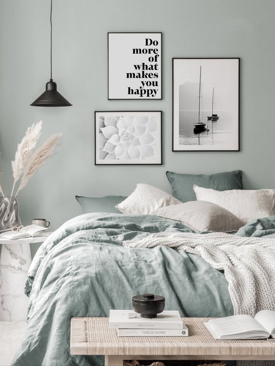 a pastel green bedroom with a bed and green and neutral bedding, a woven bench, a gallery wall of posters and a black pendant lamp
