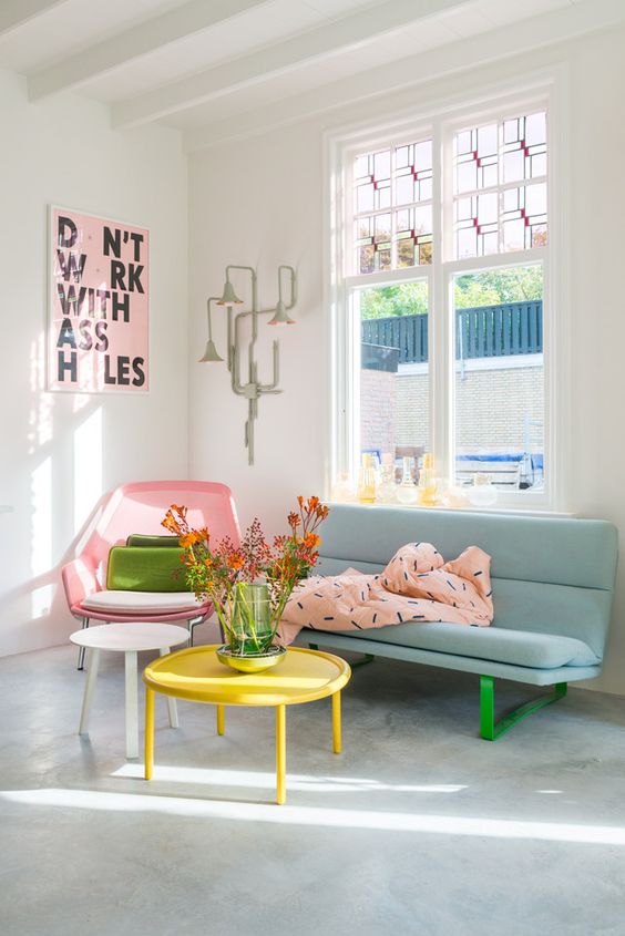 a pastel living room with a pink chair, a blue sofa with green legs, a yellow table, a pink blanket and a pink artwork