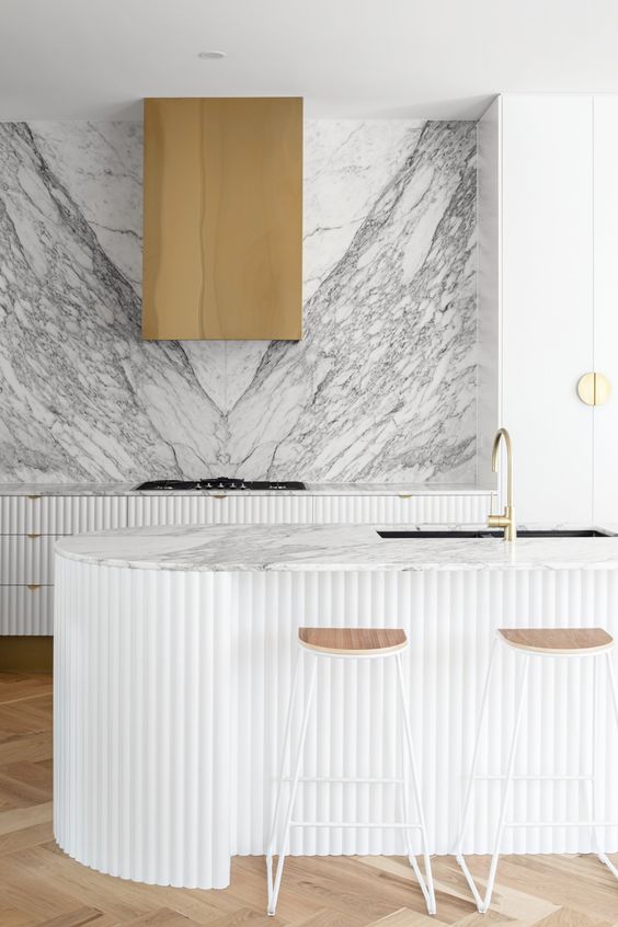 a refined contemporary kitchen with ridged cabinets and a matching curved kitchen island, a white marble backsplash plus a brass hood