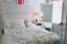 a small pastel blue bedroom with a white bed and neutral bedding, a lit up mirror, a nightstand and a gallery wall over the bed