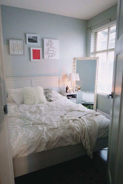 a small pastel blue bedroom with a white bed and neutral bedding, a lit up mirror, a nightstand and a gallery wall over the bed