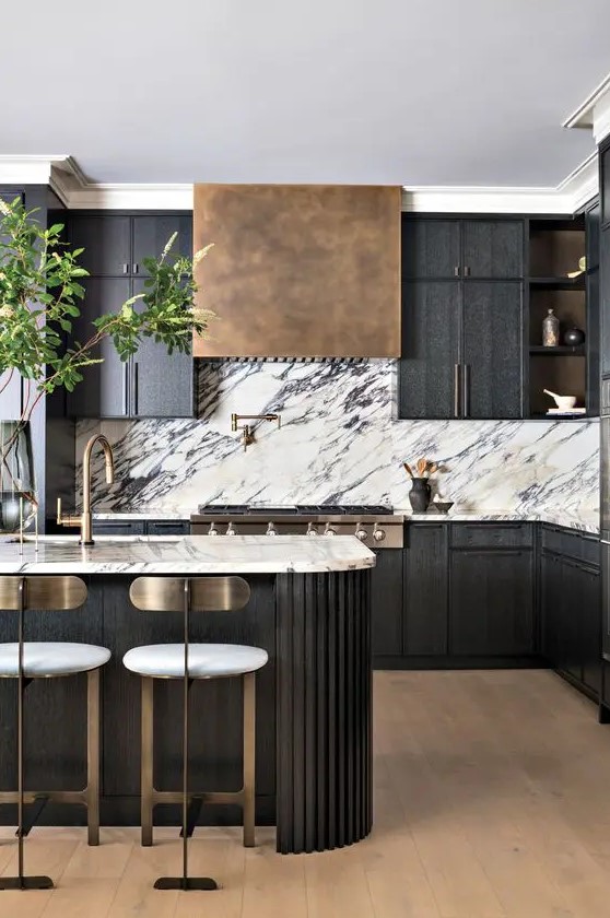 a sophisticated black kitchen with a white marble backsplash, an aged brass hood, a fluted curved kitchen island and tall stools with brass backs