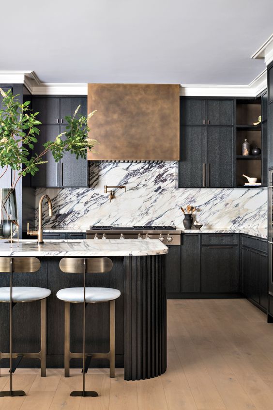 a sophisticated black kitchen with a white marble backsplash, an aged brass hood, a fluted curved kitchen island and tall stools with brass backs