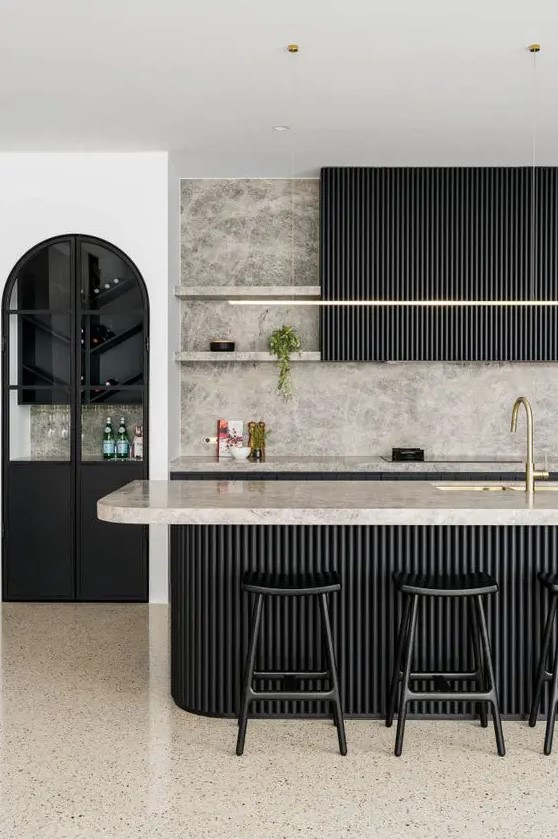 a sophisticated black kitchen with fluted cabinets and a matching kitchen island, grey marble countertops and a backsplash, gold fixtures