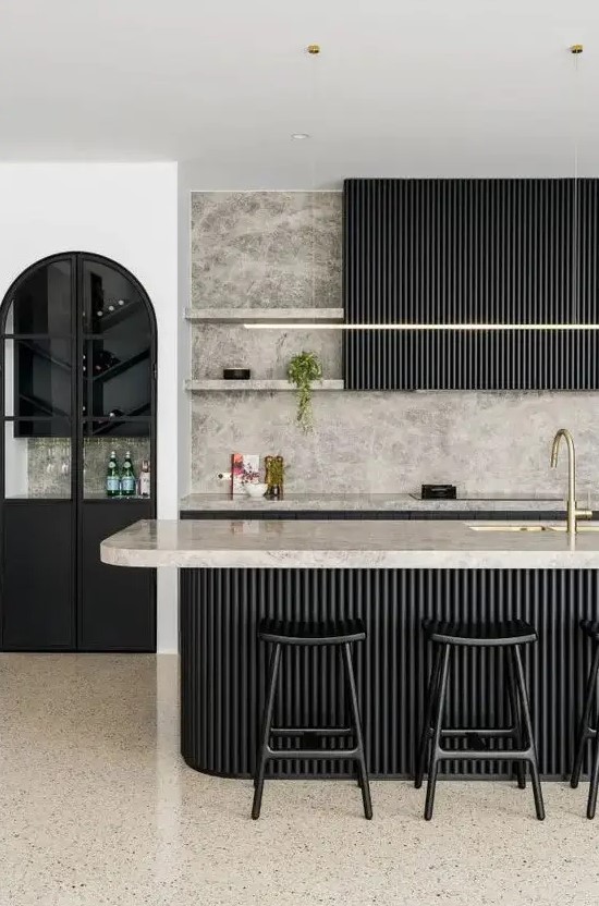 a sophisticated black kitchen with fluted cabinets and a matching kitchen island, grey marble countertops and a backsplash, gold fixtures