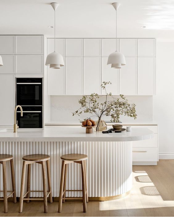 a sophisticated contemporary kitchen in white, with a white stone backsplash and countertops, a curved fluted kitchen island, pendant lamps