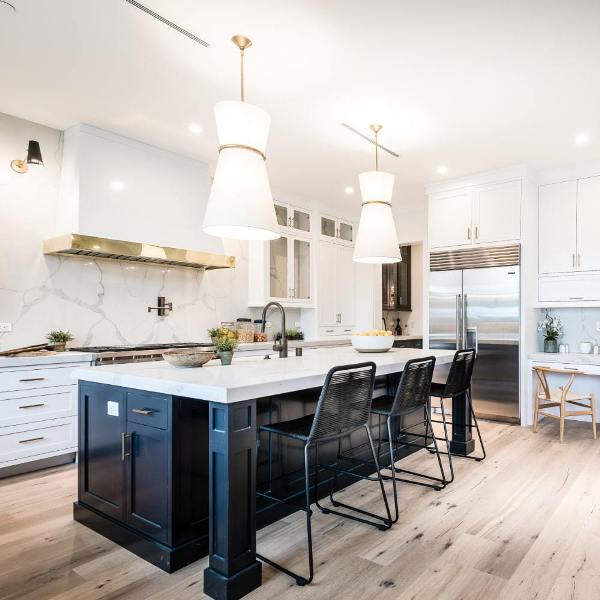 a sophisticated white kitchen with shaker cabinets, a black kitchen island, a large hood, a white quartz backsplash and matching countertops