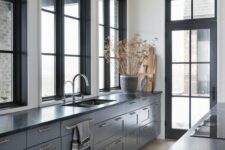 a stylish and elegant grey kitchen with shaker cbainets, black countertops, black framed windows and a door