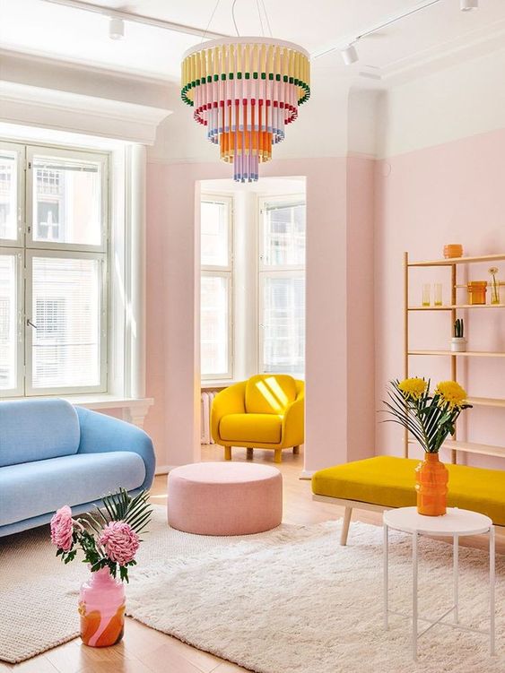 a welcoming pastel living room with pink walls, a blue sofa, a yellow chair, a yellow daybed, a pink pouf and a colorful chandelier