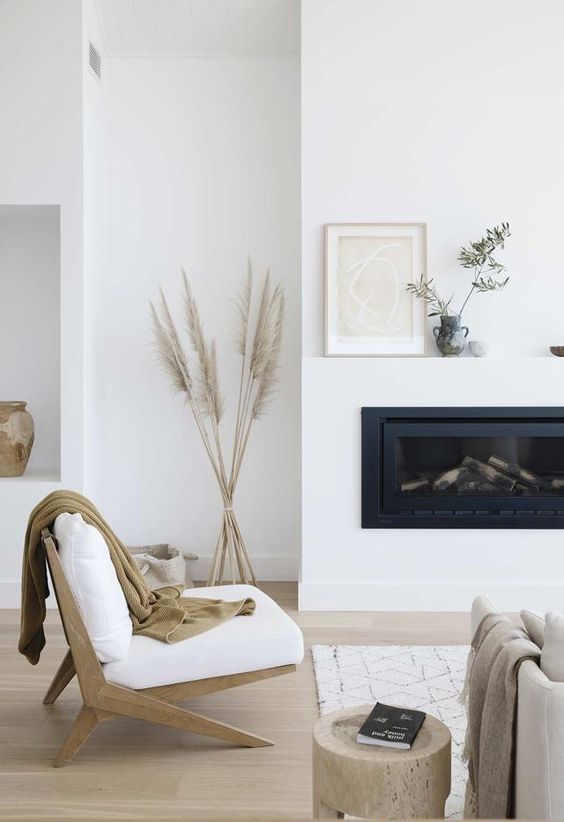 a white Scandinavian living room with a built in fireplace, a white chair,a  neutral sofa, a wooden stool and pampas grass in a vase