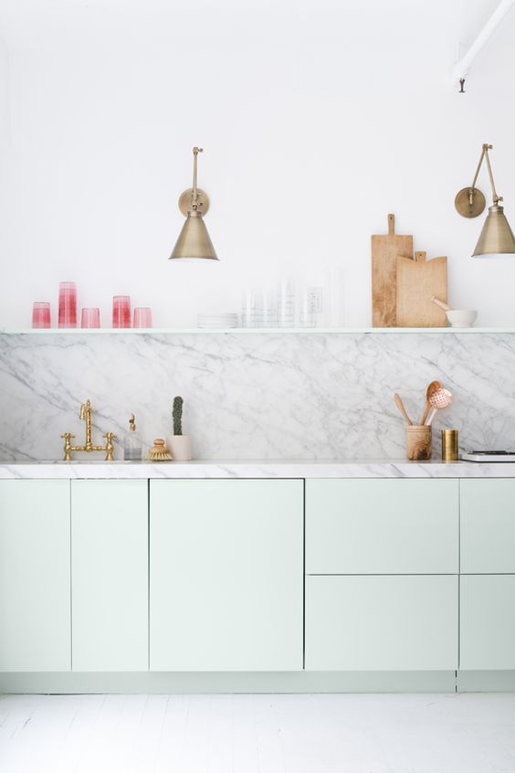 an airy pastel green kitchen with plain cabinets, a white stone countertop and a backsplash, an open shelf and brass sconces