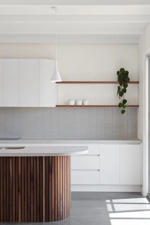 an airy white kitchen with plain cabinets and no handles, a grey skinny tile backsplash, a fluted stained kitchen island and shelves